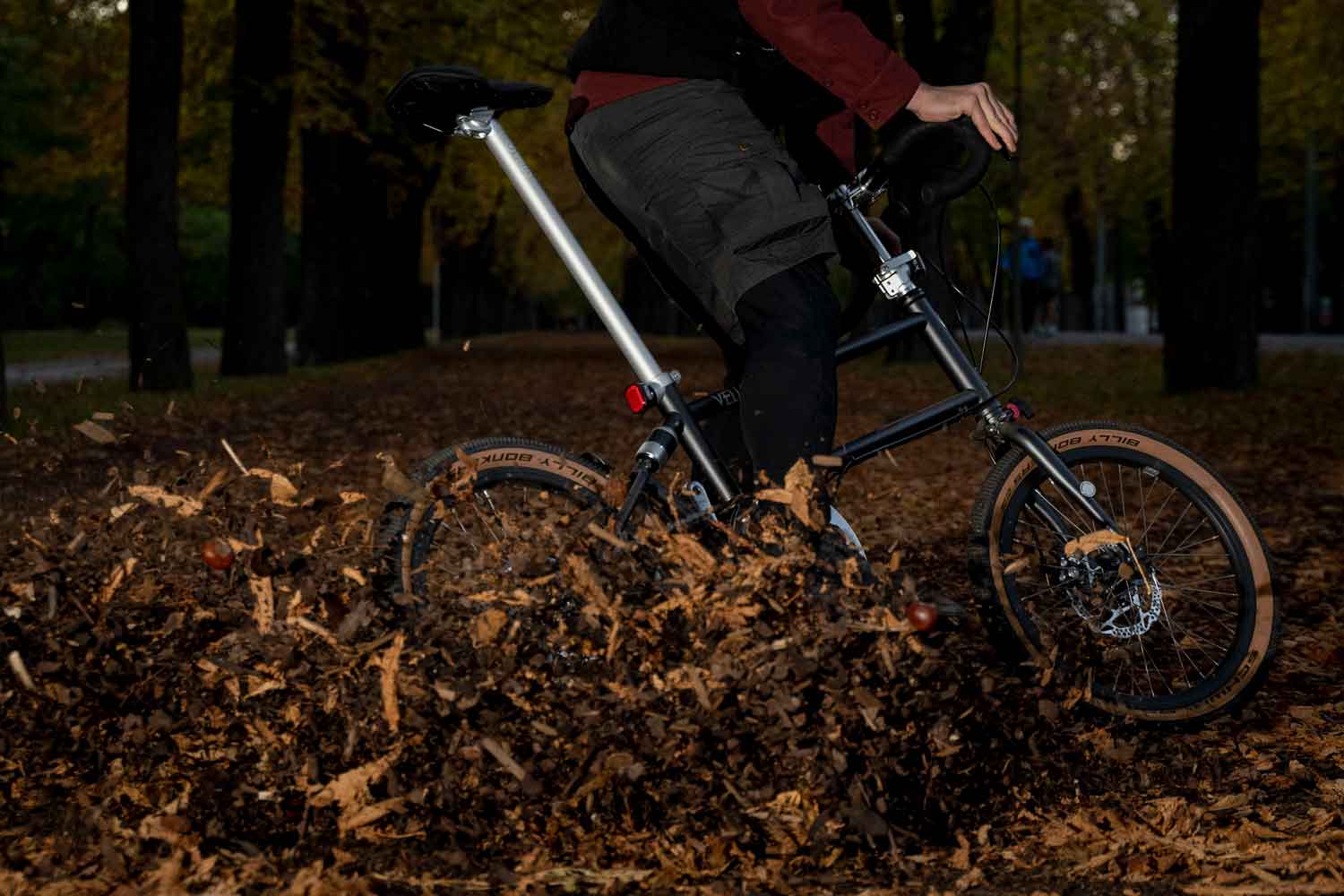 Eine Person die in einen Laubhaufen fährt und überall Blätter fliegen, mit dem VELLO Gravel Faltrad / Klapprad in der Farbe Anthrazit, mit 20 Zoll Billy Bonkers Reifen im Vordergrund.