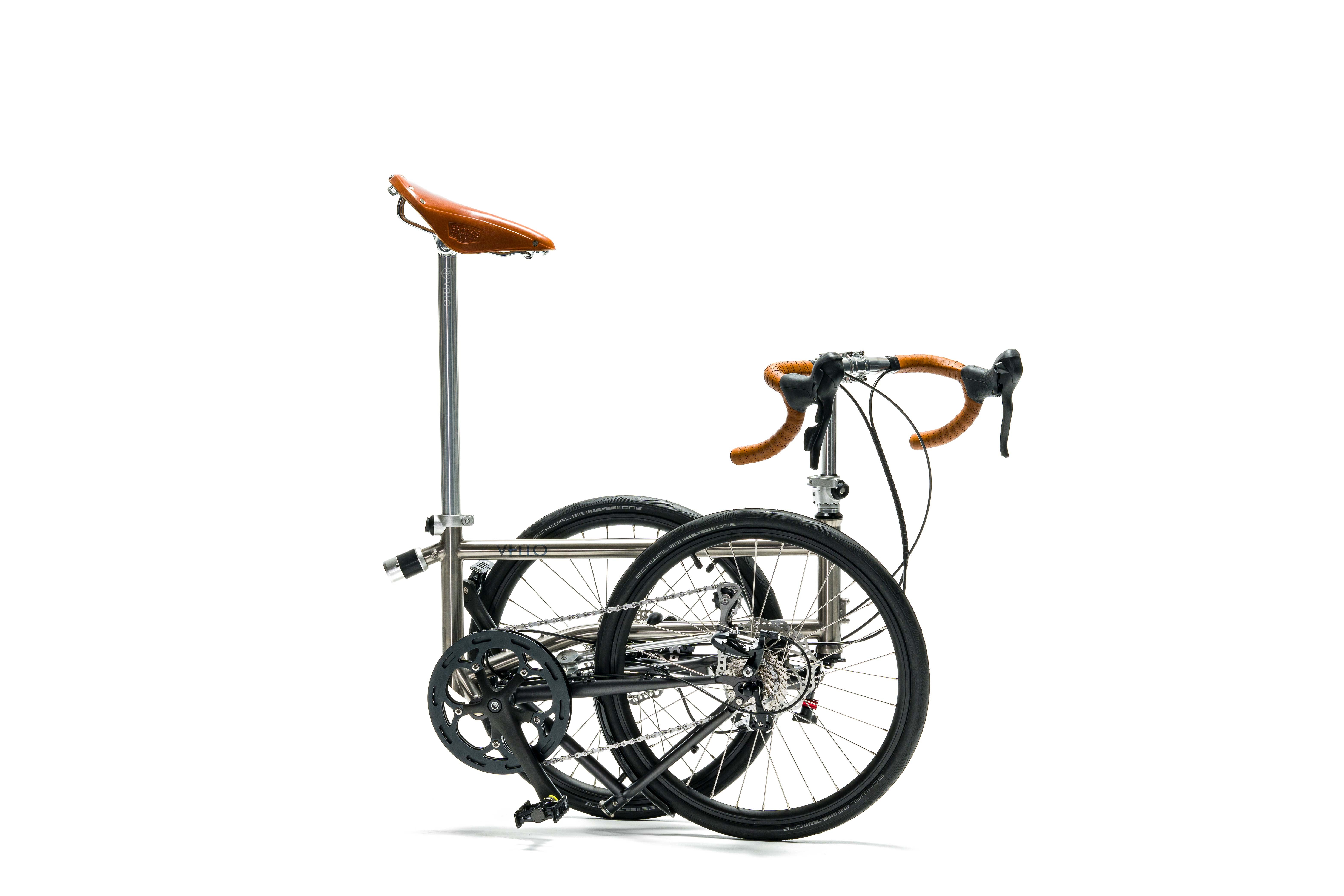 Ein gefaltetes VELLO Speedster Titan Faltrad / Klapprad ,in der Farbe gebürstetes Titan, mit 20 Zoll Schwalbe One Tube Type Performance 28-451 Reifen und den ikonischen Honigbraunen Sattel, sowie entfalteten Griffen.