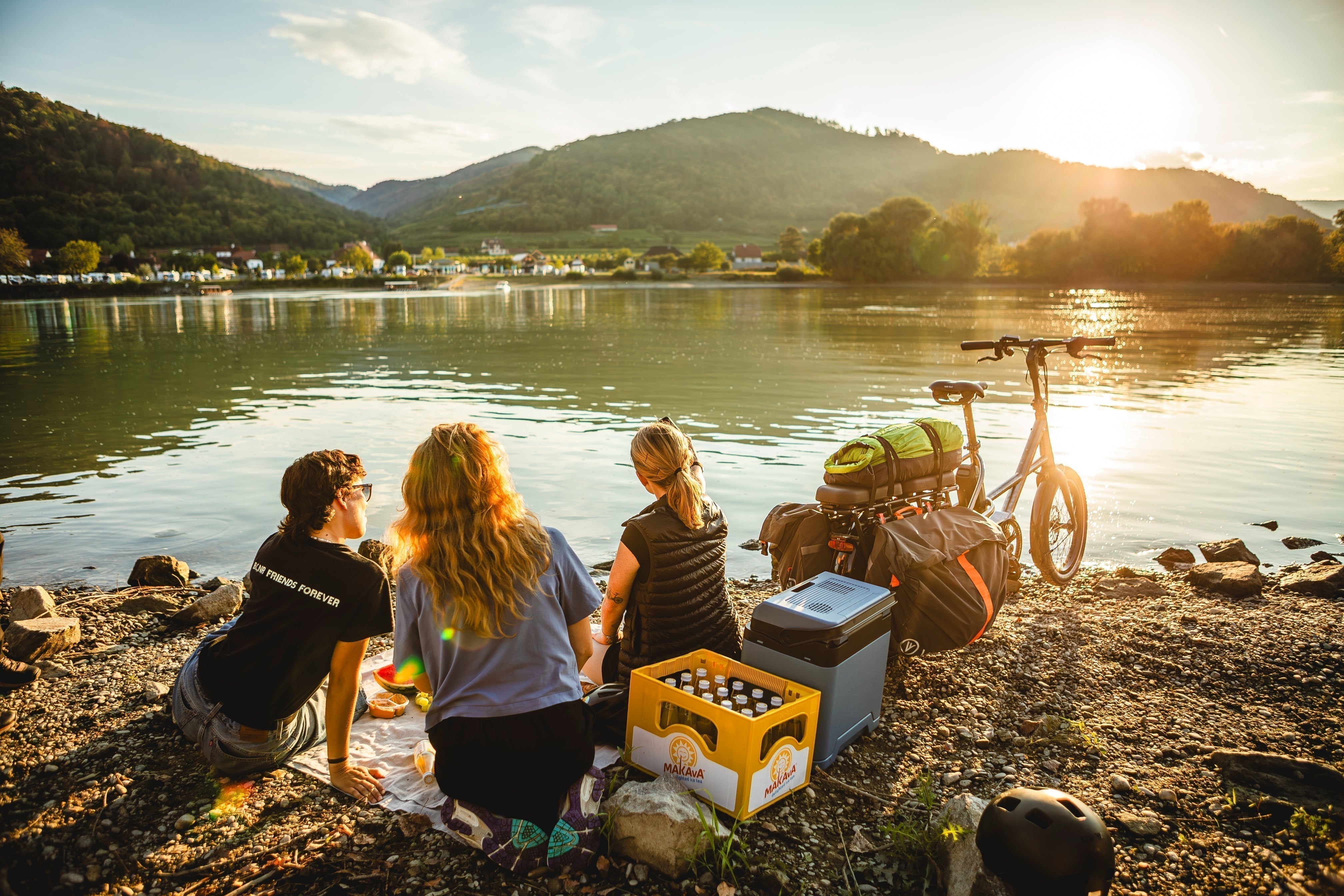 Klar — etwas präziser für Bild 4:  **Alt-Text:** Drei Personen machen bei Sonnenuntergang ein Picknick am Donauufer, neben ihnen steht ein voll beladenes VELLO SUB mit Campingausrüstung.