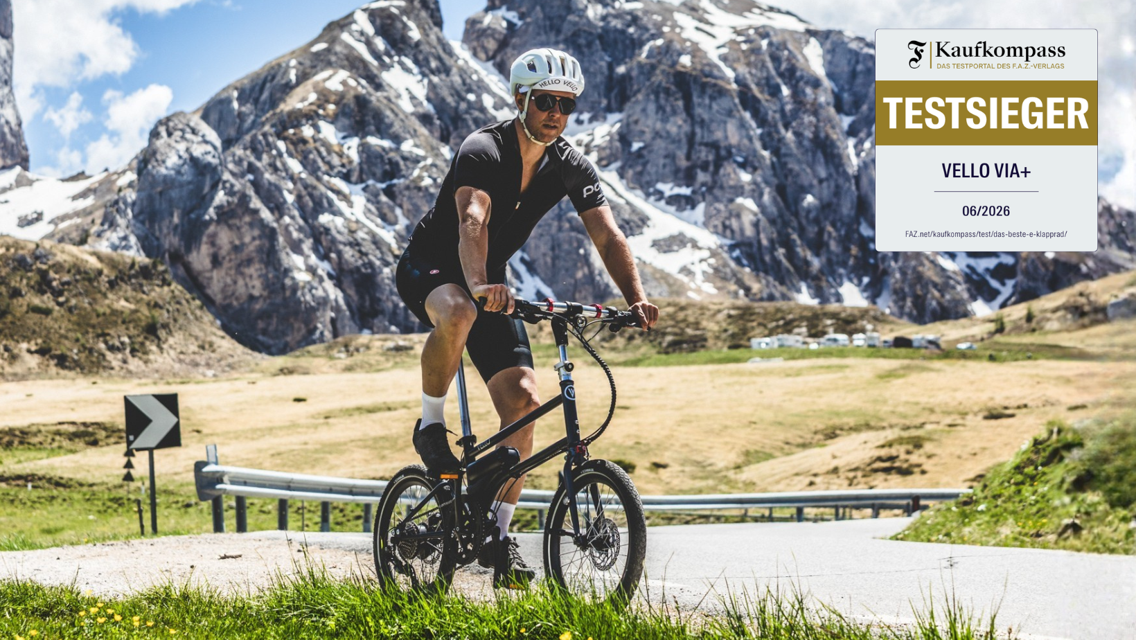Radfahrer, schwarzes VELLO VIA+, Bergstraße, schneebedeckte Alpenkulisse, Kaufkompass-Siegel „Testsieger VELLO VIA+, 06/2026“