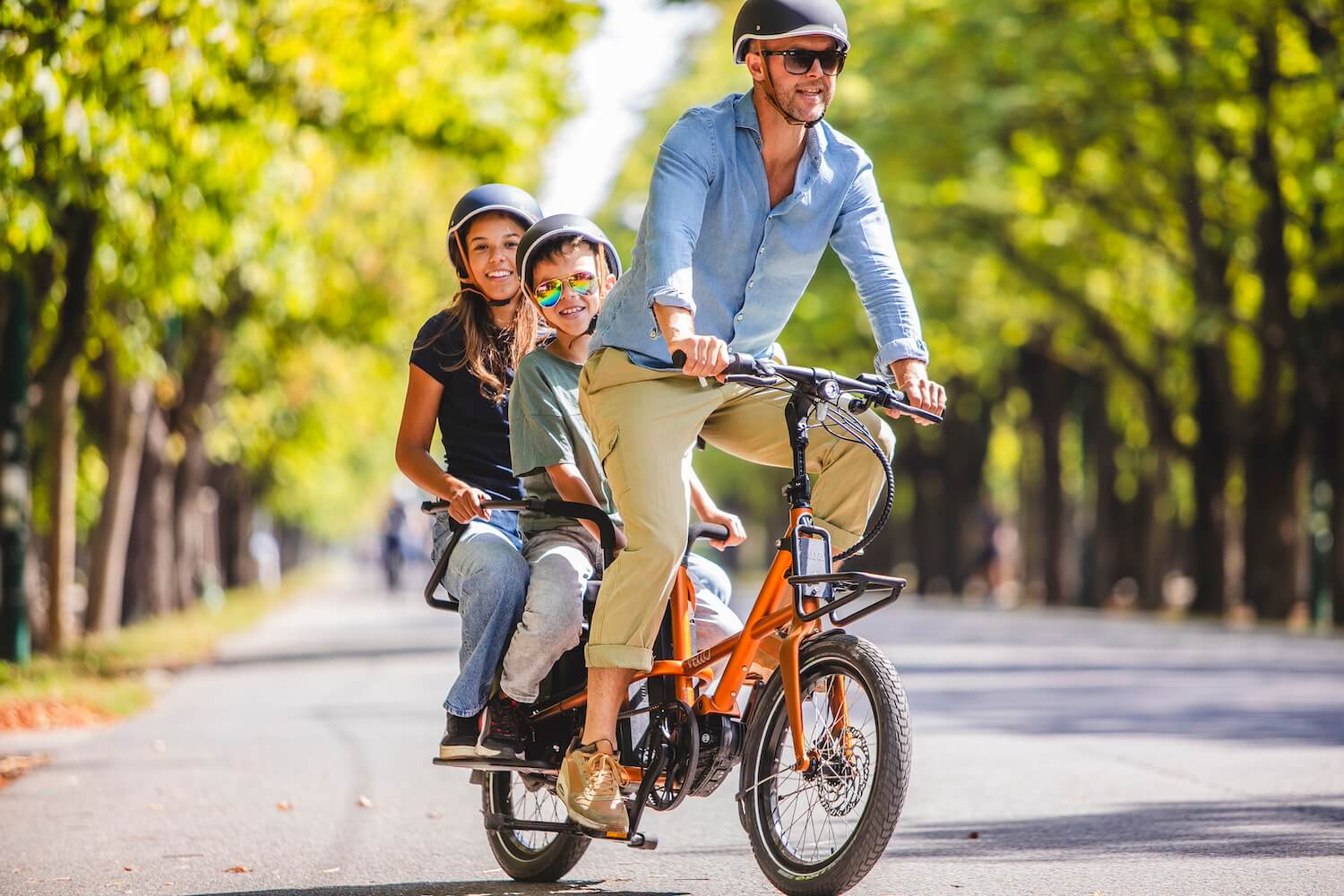 Familienausflug mit dem VELLO SUB: Ein Elternteil fährt mit zwei Kindern auf dem kompakten Longtail E-Bike durch eine grüne Parklandschaft.